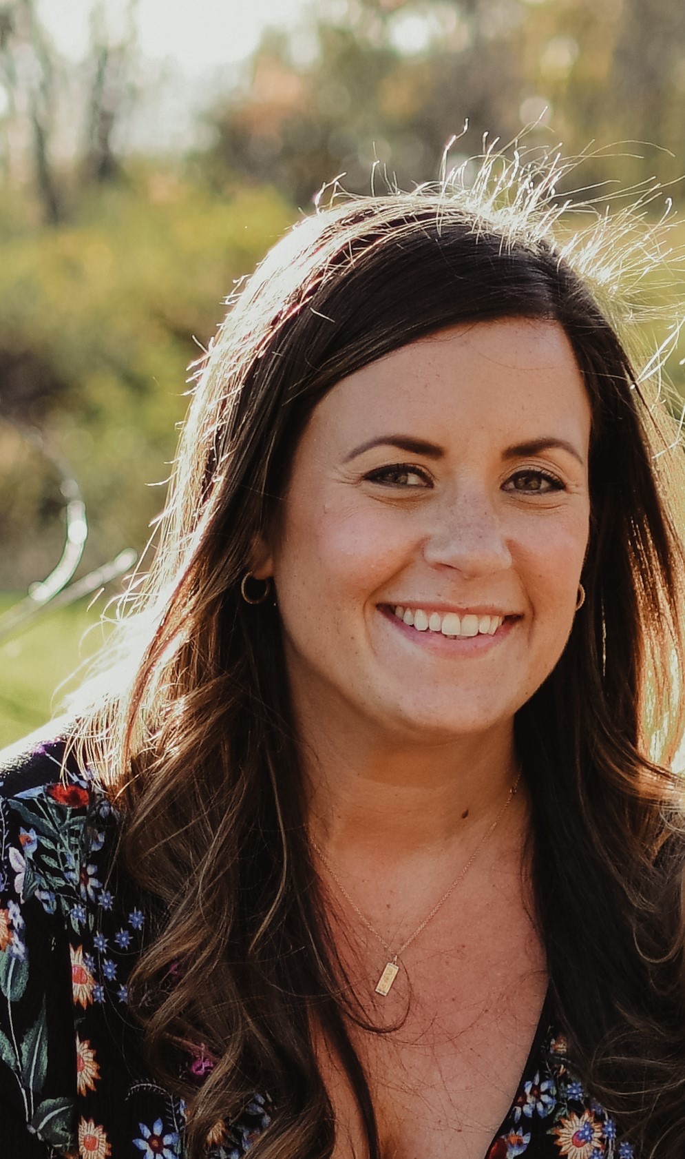 Close-up portrait of a smiling woman with shoulder-length brown hair, wearing a floral top and a necklace, with sunlit greenery in the background.