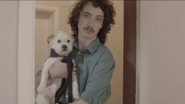 A young man with curly hair holding a small white dog while standing in a doorway. He is dressed in a blue shirt and carries a dark shoulder bag.
