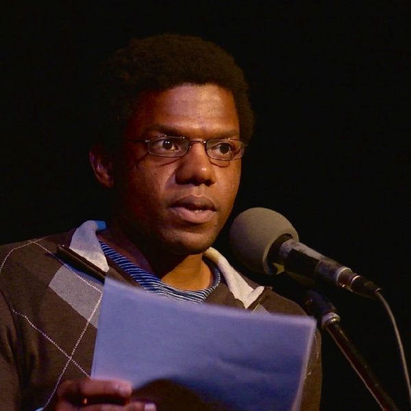 A man wearing glasses and a sweater over a collared shirt stands at a microphone, reading from papers he holds in his hands, illuminated against a dark background.