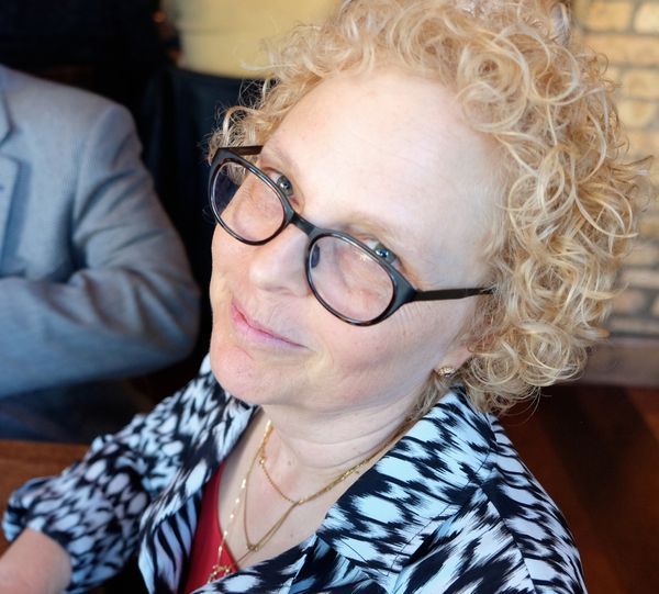 A woman with curly blonde hair and glasses smiling slightly at the camera, wearing a black and white patterned top. She is in a well-lit indoor setting.