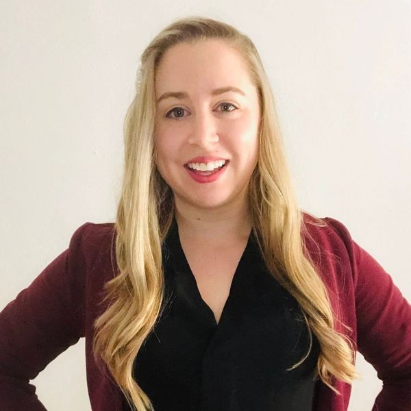 A smiling woman with long blonde hair wearing a black blouse and maroon blazer stands against a plain white background.
