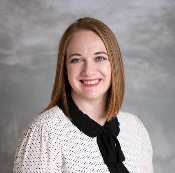 Portrait of a smiling woman with shoulder-length straight hair, wearing a white blouse with a black bow tie. The background is a textured gray.