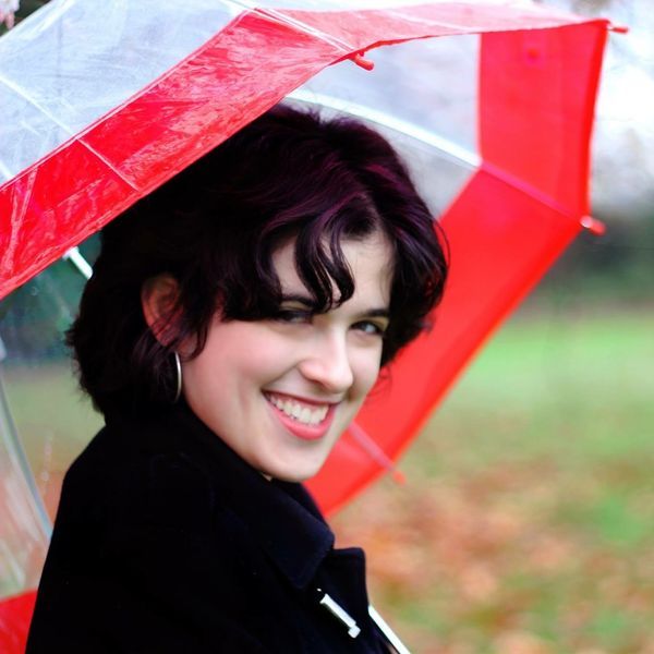 A joyful woman with dark hair smiling under a red and transparent umbrella, with a blurred green background suggesting a vibrant outdoor setting.