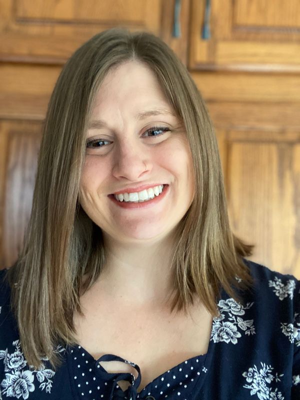A smiling woman with shoulder-length brown hair, wearing a navy blue floral blouse, stands in front of a wooden-paneled background. She has a nose ring and a cheerful expression.