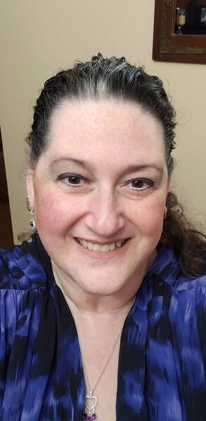 A smiling woman with dark, curly hair pulled back, wearing a blue patterned blouse and earrings, standing indoors with a framed picture in the background.