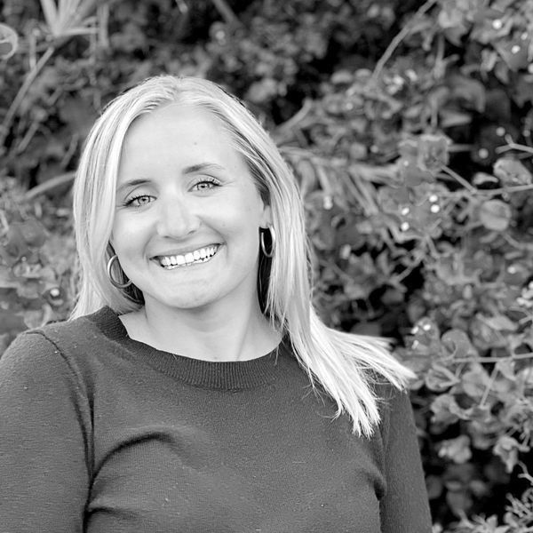 Black and white portrait of a smiling woman with shoulder-length hair wearing earrings and a dark sweater, set against a blurred background of leafy bushes.