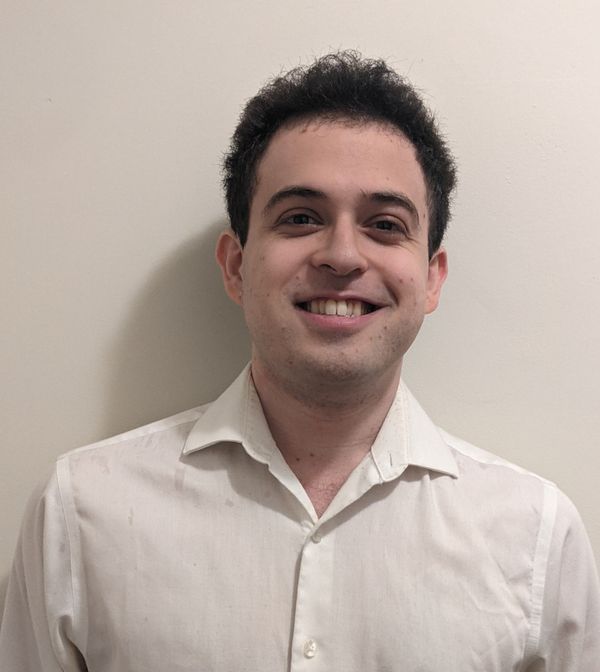 A smiling man with short dark hair, wearing a light beige shirt, stands against a plain white wall.