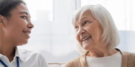 An elderly woman with white hair and a beige sweater smiles warmly while looking at a younger woman with dark hair tied back, who wears a white shirt and a name tag displaying her credentials. They sit on a sofa in a bright, sunlit room, enjoying each other's company.