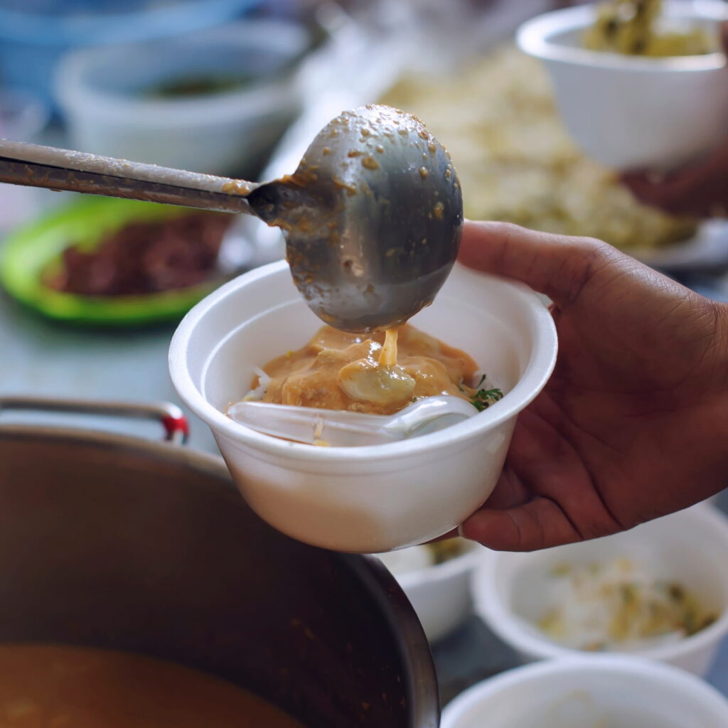 A close-up of a hand holding a white bowl while another person ladles hot soup or curry over noodles, preparing a meal. Other bowls and ingredients are visible in the background.