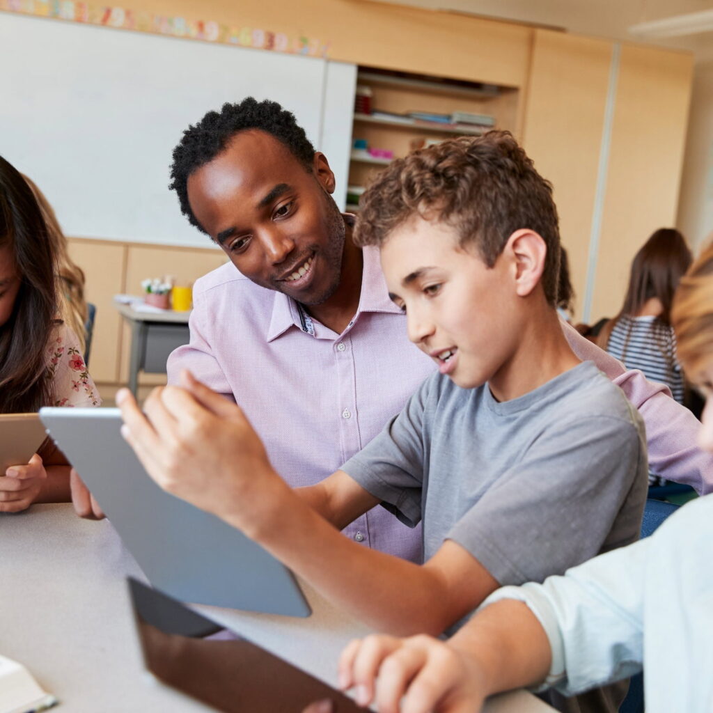 A teacher sits at a table with students in a classroom, smiling and helping a boy who is using a tablet. Other students nearby are also engaged with their tablets.