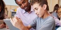 A teacher sits at a table with students in a classroom, smiling and helping a boy who is using a tablet. Other students nearby are also engaged with their tablets.