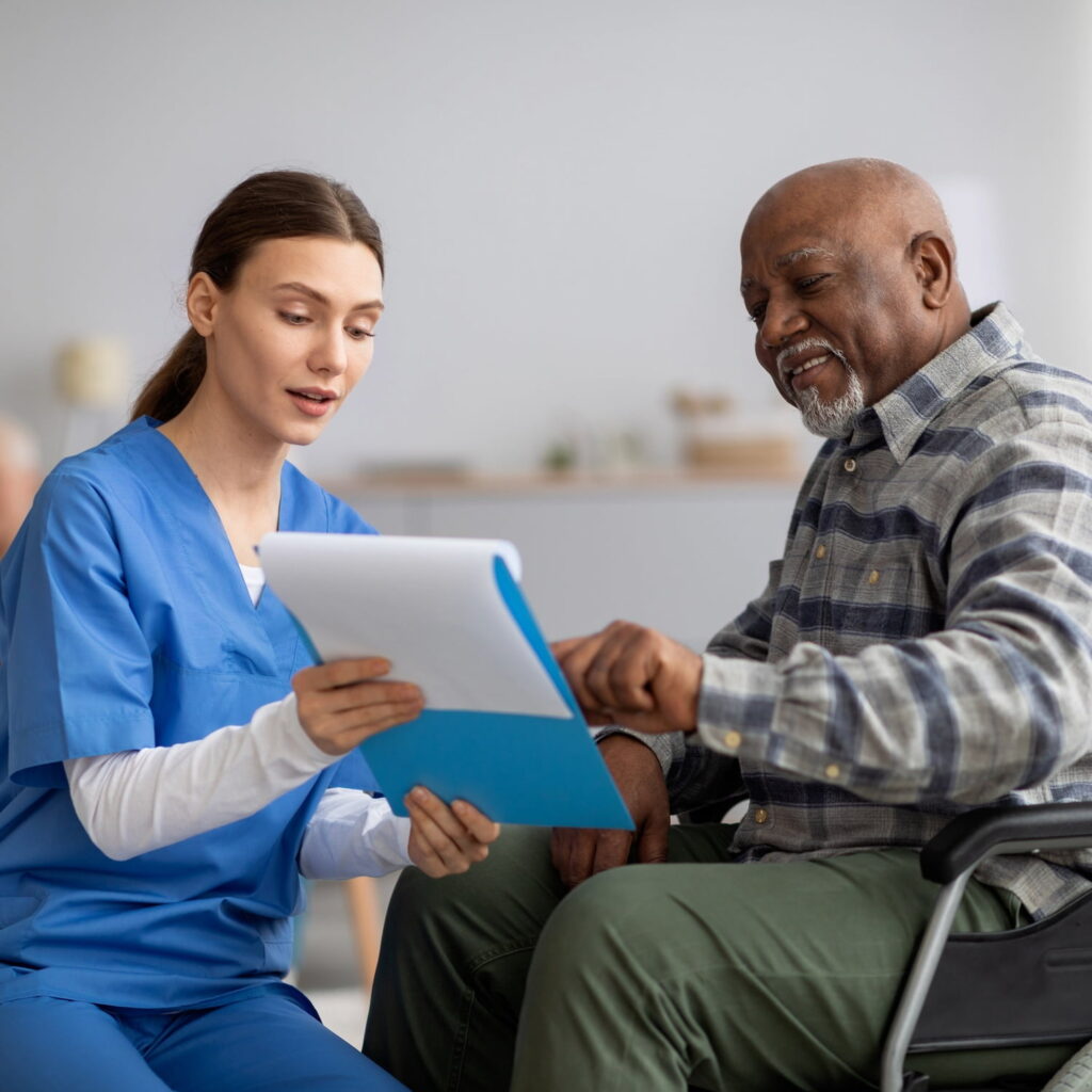 A nurse in blue scrubs shows a clipboard with documents to an older man in a wheelchair. They are both smiling and discussing the paperwork in a bright, comfortable room.