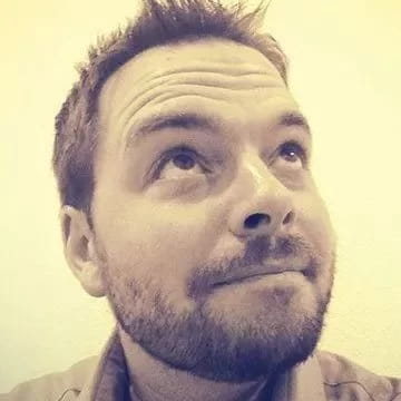 A bearded man with short hair looks upward with a slight smile. The image has a sepia tone, and the background is plain and light-colored.